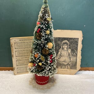 May include: A miniature Christmas tree with green needles, white flocking, and red, yellow, and brown decorations. The tree is in a red pot on a lace doily. Vintage sheet music and a sepia-toned portrait are in the background.