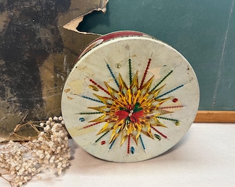 May include: A vintage round tin box with a white background and a colorful starburst design. The starburst is made of gold, red, green, and blue paper and has a beaded border.