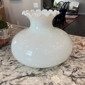 May include: A white glass lamp shade with a ruffled edge. The shade has a round base and a flared top. The glass is opaque and has a glossy finish. The lamp shade is sitting on a granite countertop.
