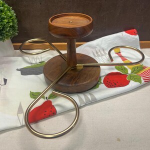 May include: A wooden candle holder with a circular base and top, connected by a central stem. Three curved, gold-colored metal arms extend from the base. The holder sits on a white patterned cloth with red and green fruit designs.