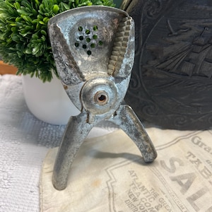 May include: A vintage, silver-toned citrus juicer with a hinged design. The juicer has a perforated bowl and a ridged reamer. The background includes a white cloth with text and a green plant in a white pot.