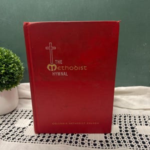 May include: A red hardcover book titled The Methodist Hymnal with a white cross emblem and gold lettering on the front cover. The text Columbia Methodist Church is printed in small gold letters at the bottom of the front cover.