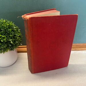 May include: A vintage, red hardcover book with a faded cover and worn edges. The book is standing upright, revealing its aged, cream-colored pages. A small, green, artificial plant in a white pot is in the background.