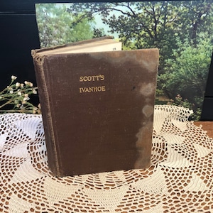 May include: A vintage, brown leather-bound book with the title "SCOTT'S IVANHOE" embossed in gold lettering. The book is sitting on a white crocheted doily.