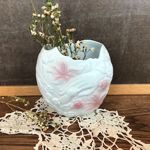 May include: White ceramic vase with a unique, organic shape, adorned with embossed floral and hand motifs. The vase features pink accents on the flowers and holds dried flowers. It sits on a lace doily, creating a vintage aesthetic.