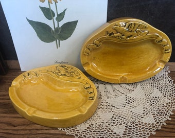 May include: Two vintage, oval-shaped ashtrays in a mustard yellow hue. Each ashtray features a raised relief design of an eagle with outstretched wings and decorative scrollwork. The ashtrays are made of ceramic and have a glossy finish.