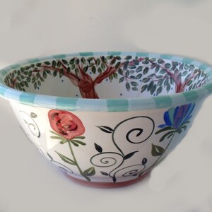 May include: A large white ceramic bowl with a hand-painted design featuring a green olive tree, red flowers, and blue flowers. The bowl has a light blue and white striped rim.