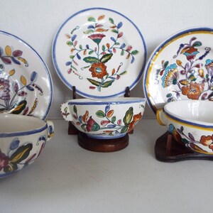 May include: Set of four vintage ceramic bowls and plates with floral designs in shades of blue, yellow, green, red, and white. The bowls have two handles and are on wooden stands.