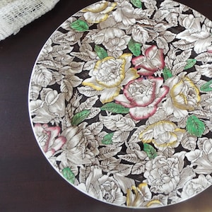May include: A round ceramic plate with a floral pattern. The plate features a black background with white, yellow, and pink roses and green leaves.
