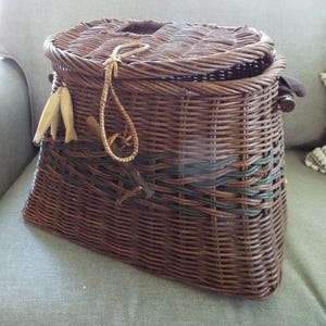 May include: A dark brown woven wicker basket with a hinged lid and a loop closure. The basket features a horizontal band of dark green woven accents and two small, light-colored wooden fish-shaped charms hanging from the front side.