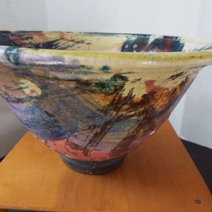 May include: A large, ceramic bowl with a wide, flared rim and a rounded base. The bowl is decorated with abstract brushstrokes in shades of blue, green, yellow, orange, and red.