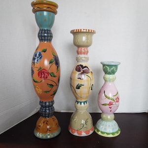 May include: Set of three hand-painted candle holders with floral designs. The tallest holder is orange with blue and white accents, the middle holder is yellow with green and white accents, and the shortest holder is pink with green and white accents.