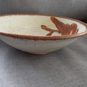 May include: A shallow ceramic bowl with a cream-colored base and a wide, dark brown rim. The interior features a hand-painted, abstract brown brushstroke design that extends from the side toward the center, with subtle concentric circular grooves.