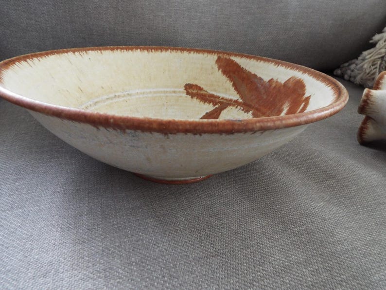 May include: A shallow ceramic bowl with a cream-colored base and a wide, dark brown rim. The interior features a hand-painted, abstract brown brushstroke design that extends from the side toward the center, with subtle concentric circular grooves.