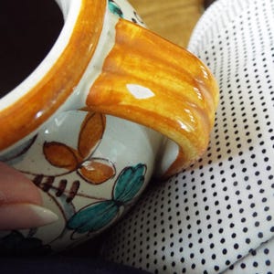 May include: A ceramic mug with a glossy cream-colored base, featuring hand-painted floral motifs in shades of teal and orange. The mug has a thick, curved handle with a warm amber-colored glaze and a dark interior, showcasing a smooth finish.