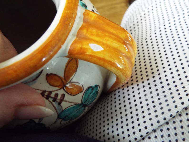 May include: A ceramic mug with a glossy cream-colored base, featuring hand-painted floral motifs in shades of teal and orange. The mug has a thick, curved handle with a warm amber-colored glaze and a dark interior, showcasing a smooth finish.