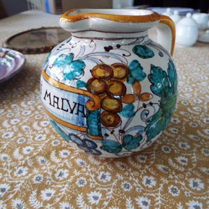 May include: A round ceramic pitcher with a cream-colored glaze, featuring hand-painted green leaves and clusters of brown grapes. A yellow banner across the front displays the text MALVA in black letters. The pitcher has a single handle.