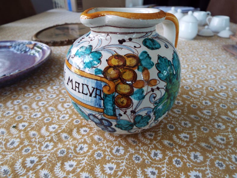 May include: A round ceramic pitcher with a cream-colored glaze, featuring hand-painted green leaves and clusters of brown grapes. A yellow banner across the front displays the text MALVA in black letters. The pitcher has a single handle.