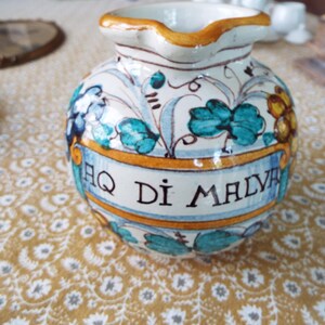 May include: A round ceramic apothecary jar with a spout, featuring a hand-painted floral pattern in teal, yellow, and blue. A central banner across the front displays the black text "AQ DI MALVA" framed by yellow borders, with a glossy glazed finish.