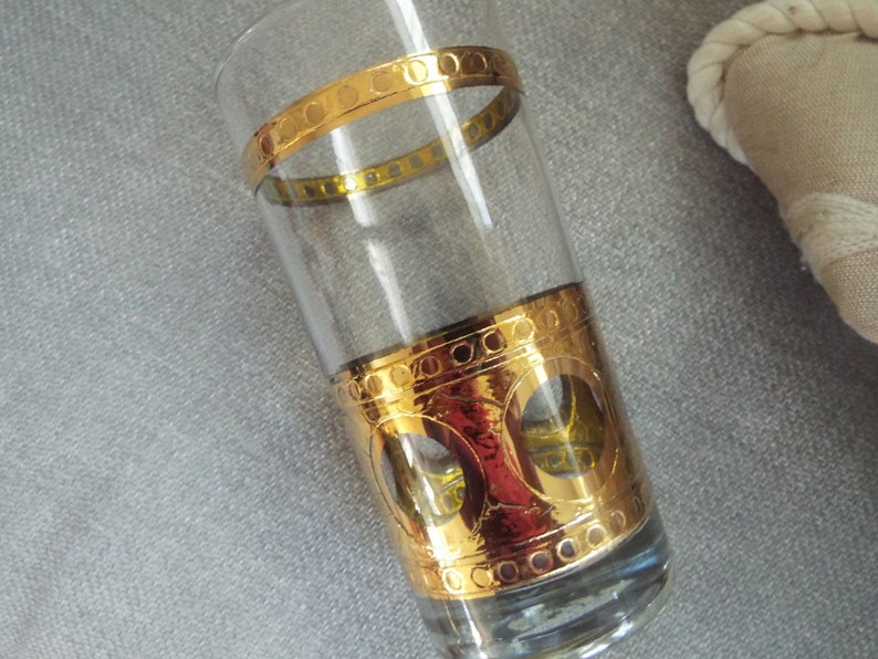 May include: A clear glass tumbler with a gold band and a gold design around the bottom. The design features circles and a pattern of small dots.