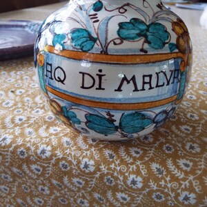 May include: A round ceramic apothecary jar featuring a cream-colored base with hand-painted teal leaves and yellow floral accents. A central banner displays the black text AQ DI MALVA. The vessel has a glossy finish and a wide, bulbous body shape.
