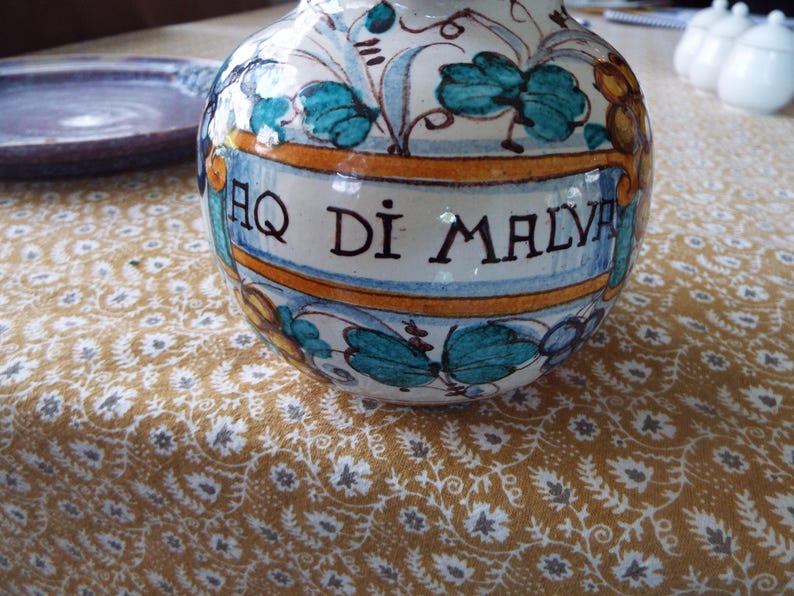 May include: A round ceramic apothecary jar featuring a cream-colored base with hand-painted teal leaves and yellow floral accents. A central banner displays the black text AQ DI MALVA. The vessel has a glossy finish and a wide, bulbous body shape.