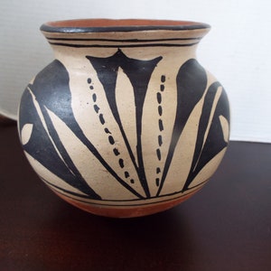 May include: A small, round, terracotta pot with a black and white geometric design. The pot has a wide mouth and a rounded base.