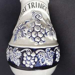 May include: A white ceramic pitcher with blue and white grape designs. The pitcher has the words "Art Trinkt" written on the side.