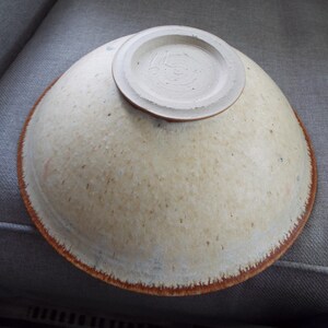 May include: A ceramic bowl shown upside down, featuring a speckled cream and off-white glaze with a distinct reddish-brown rim. The base of the bowl is circular, raised, and unglazed, displaying a textured surface with visible potter's marks.