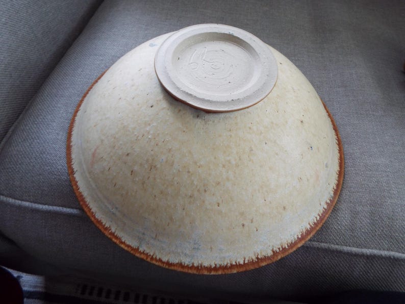 May include: A ceramic bowl shown upside down, featuring a speckled cream and off-white glaze with a distinct reddish-brown rim. The base of the bowl is circular, raised, and unglazed, displaying a textured surface with visible potter's marks.
