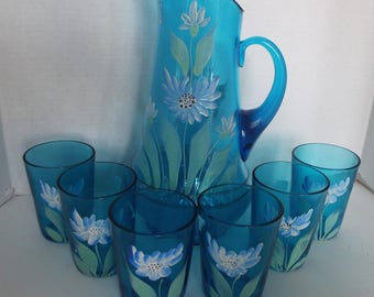 May include: A set of seven turquoise blue glass tumblers and a pitcher with hand-painted white flowers. The pitcher has a wide mouth and a handle.