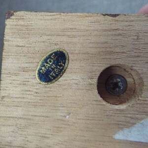 May include: Close-up of a wooden base with a blue and gold sticker that reads "Made in Italy". There is a screw in the center of the base.
