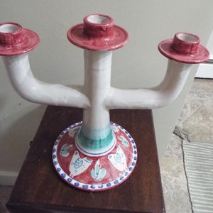 May include: A white ceramic three-arm candelabra with red and green painted details. The base features a circular design with stylized birds and leaves.