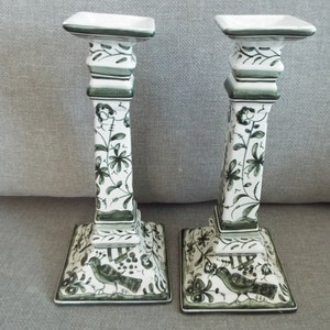 May include: A pair of white ceramic candle holders with green floral and bird designs. The holders have a square base and a tall, rectangular column.