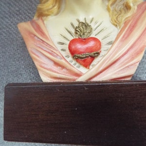 May include: A bust of the Sacred Heart of Mary, a religious figurine with a heart on her chest. The heart is red with a gold crown of thorns and a gold rope around it. The bust is made of white painted plaster and is mounted on a dark wood base.