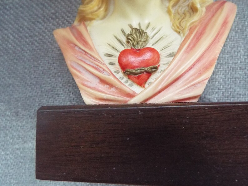 May include: A bust of the Sacred Heart of Mary, a religious figurine with a heart on her chest. The heart is red with a gold crown of thorns and a gold rope around it. The bust is made of white painted plaster and is mounted on a dark wood base.