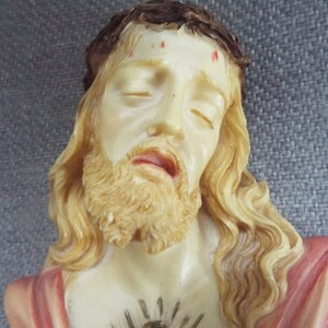 May include: A white plaster bust of Jesus Christ with a crown of thorns, long flowing hair, and a beard. He is wearing a red robe and has a heart with a crown of thorns on his chest.