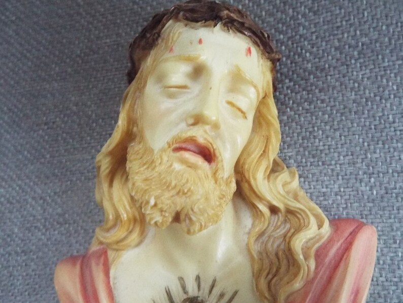 May include: A white plaster bust of Jesus Christ with a crown of thorns, long flowing hair, and a beard. He is wearing a red robe and has a heart with a crown of thorns on his chest.