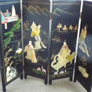 May include: Black wooden folding screen with four panels. Each panel features a detailed, raised relief design with figures in traditional clothing, landscapes, and floral elements. The artwork is accented with gold and other colors.