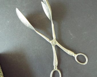 May include: Silver-toned serving tongs with ornate floral detailing. The tongs have a scissor-like design with rounded handles and a circular opening at the end. The tongs are open, ready to serve.