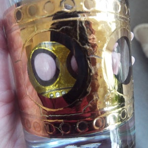May include: A gold-rimmed glass shot glass with a black and yellow design featuring pink circles. The glass has a textured surface.