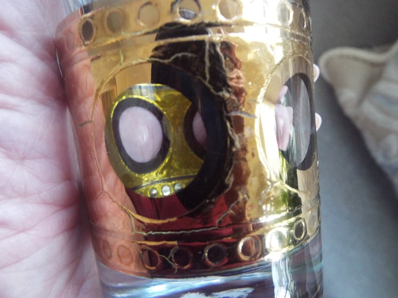 May include: A gold-rimmed glass shot glass with a black and yellow design featuring pink circles. The glass has a textured surface.