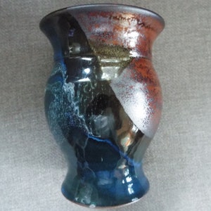 May include: A handcrafted ceramic vase with a flared rim and a rounded base. The vase features a blend of colors, including blue, black, silver, and orange. The surface has a textured appearance, adding to its unique aesthetic.