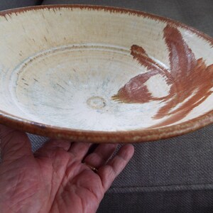 May include: A shallow ceramic bowl with a cream-colored interior and a wide, brown-glazed rim. The interior features a hand-painted, abstract brown brushstroke design that extends from the center toward the edge, with a smooth, glossy finish.