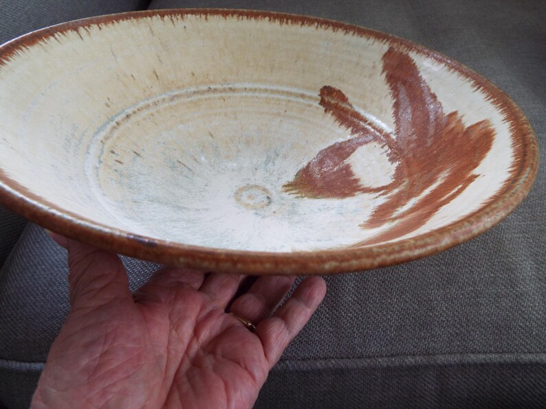 May include: A shallow ceramic bowl with a cream-colored interior and a wide, brown-glazed rim. The interior features a hand-painted, abstract brown brushstroke design that extends from the center toward the edge, with a smooth, glossy finish.
