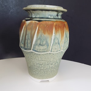 May include: A ceramic vase with a light green glaze and brown accents. The vase has a rounded body with a flared top and a wide base. It has a textured surface and a unique design.