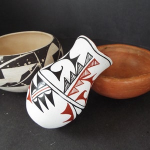 May include: Three Native American pottery pieces, a small bowl with a black and white geometric design, a larger bowl with a plain brown finish, and a small vase with a white background and black and red geometric designs.