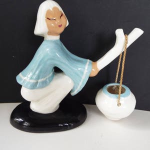 May include: A ceramic figurine of a woman in a blue and white dress carrying a white ceramic pot with a rope handle. The figurine is standing on a black base.