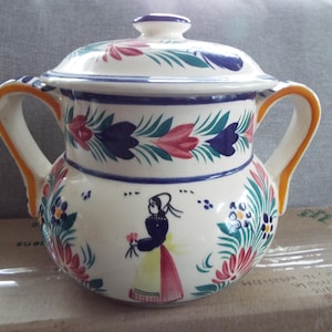 May include: A white ceramic sugar bowl with a lid, featuring a hand-painted floral design in shades of blue, green, pink, and yellow. The bowl has two handles and a stylized woman figure in the center.
