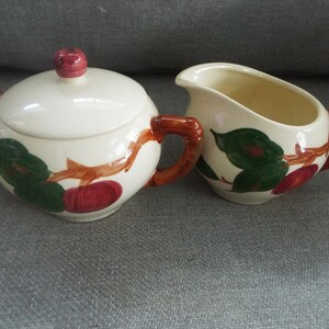 May include: A vintage cream-colored sugar bowl and creamer set with a brown twig and red apple design. The set has brown handles.